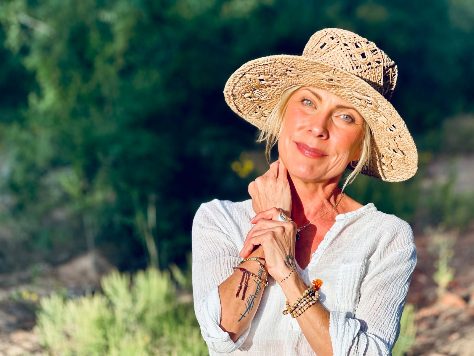 Eva Maurice outdoors in a straw hat and white shirt, hands clasped near her chin, smiling softly with green hillside behind her